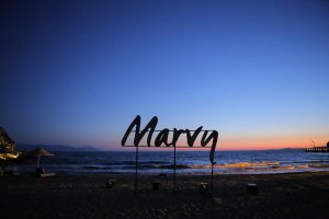 "Blick auf das 'Marvy'-Schild am Strand bei Sonnenuntergang. Der Himmel ist in orange, lila und pink getaucht. Im Hintergrund ist das Meer zu sehen."